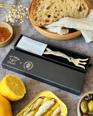 Tabletop stilll life image of the Sardine Fork shown in its presentation box surrounded by a bowl of bread, a lemon, and an open tin og sardines.